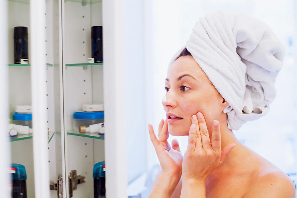 Woman with bath towel on her head, touching her skin in front of the mirror