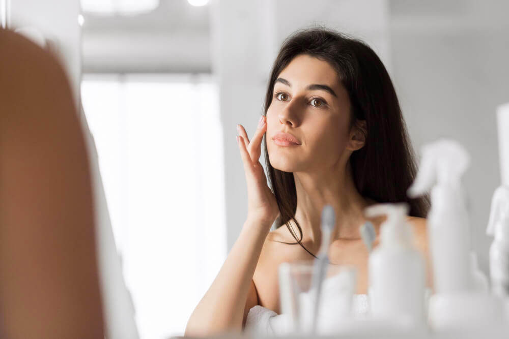 Woman applying skincare on face in front of bathroom mirror